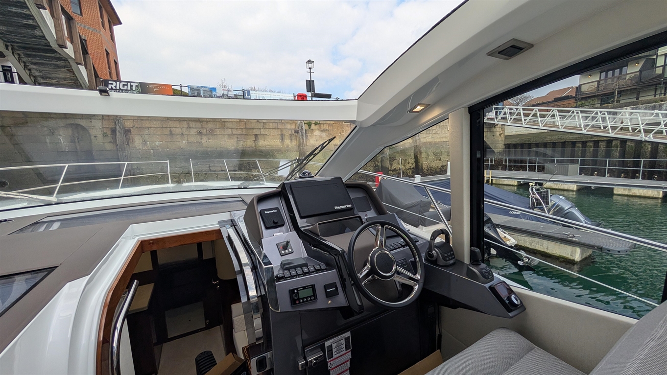 Boat cockpit view featuring a steering wheel, navigation controls, and a display screen, overlooking a marina with boats.
